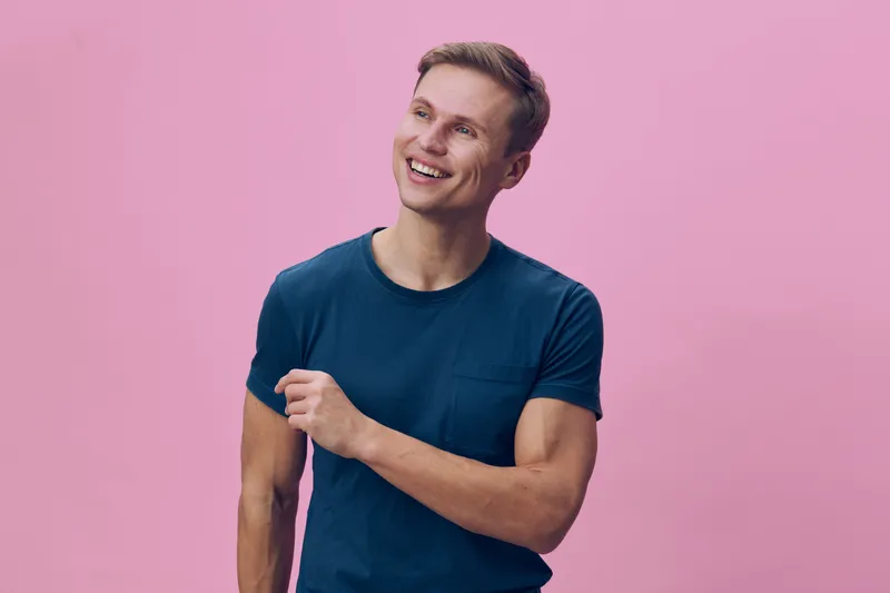 Smiling young man with short hair and casual blue t-shirt, looking happy and cheerful, standing against a plain pink background. Portrait shot capturing positive emotions and youthful energy, studio l