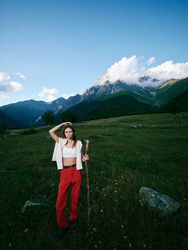 Hiking woman stands on a grassy alpine meadow with a trekking staff, red pants and white top, mountains in the background and a clear sky for a serene outdoor adventure