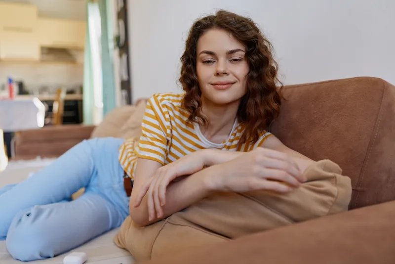 Relaxed woman lounges on a sofa at home, wearing a striped tee and jeans, resting with a soft pillow, smiling softly, surrounded by warm light, capturing casual comfort and everyday lifestyle in a
