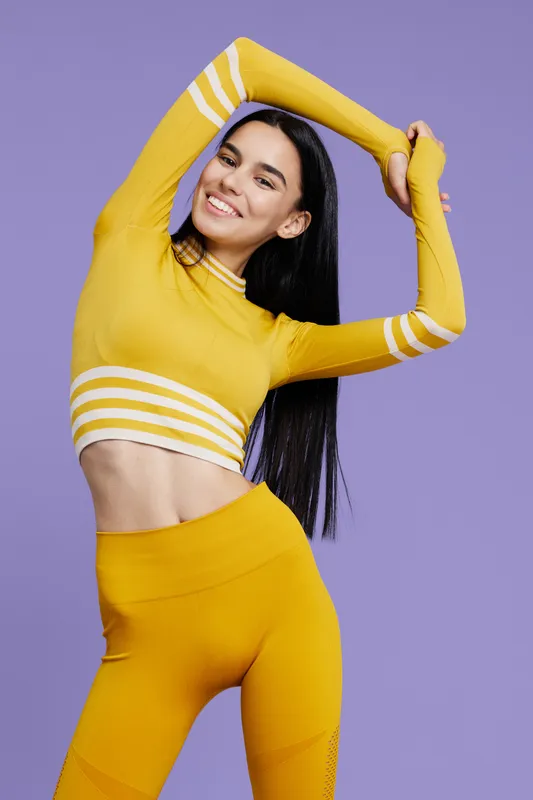 Woman sport fitness stretching in yellow activewear, smiling with long black hair, showing flexibility and energy on purple background, healthy lifestyle concept.
