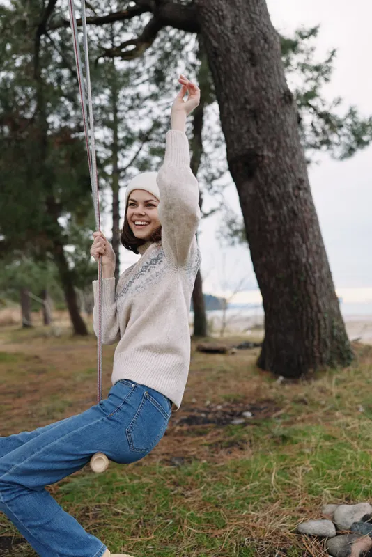 Woman on a swing outdoors in a park setting, smiling as she enjoys a carefree moment among trees, grass, and a light breeze under a cloudy sky.