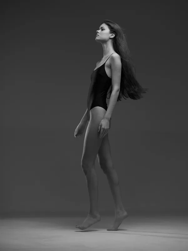 Black and white side profile of a slender woman in a leotard with long hair walking gracefully on gray background.
