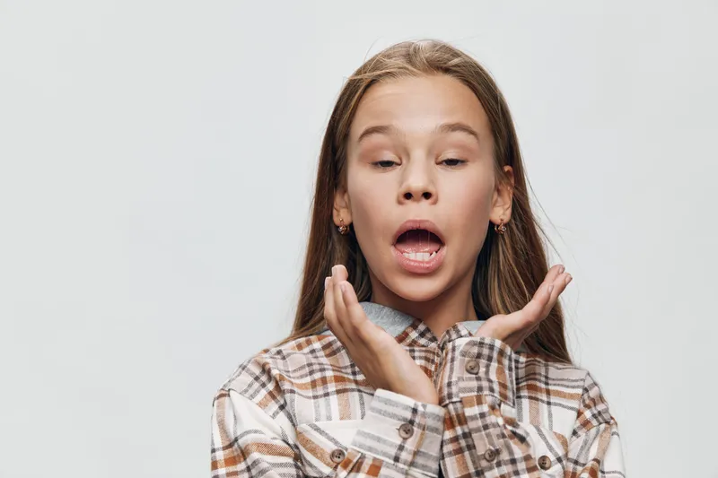Girl sneezing with closed eyes and open mouth in plaid shirt against white background for health and illness concept. Child showing allergy or cold symptoms indoors.