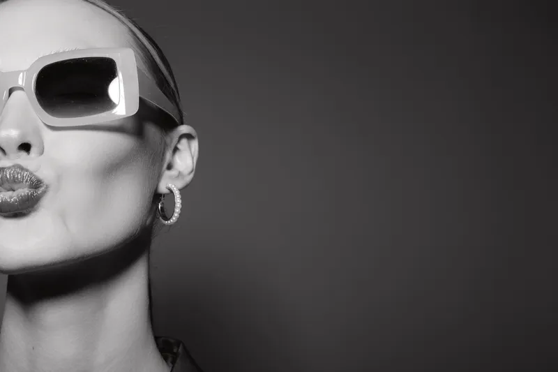 Close-up of a young fair-skinned Caucasian woman wearing stylish sunglasses sending a playful kiss. Monochrome studio portrait with smooth lighting and minimalistic background.