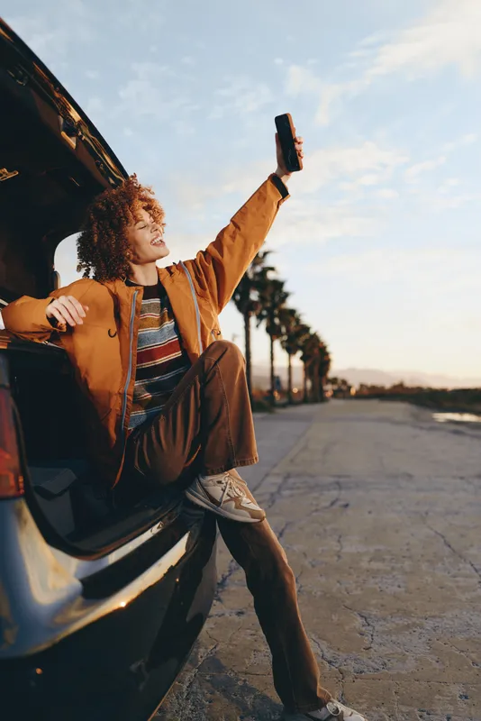Smiling woman with curly hair using smartphone near car on a city street, lifestyle travel moment outdoors, golden sunlight, casual fashion, relaxed pose