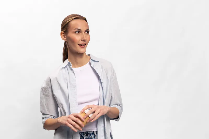 Young woman with light skin and braided hair wearing casual white shirt and jeans holds a small jar with cream. She looks away with a slight smile on a clean white background.