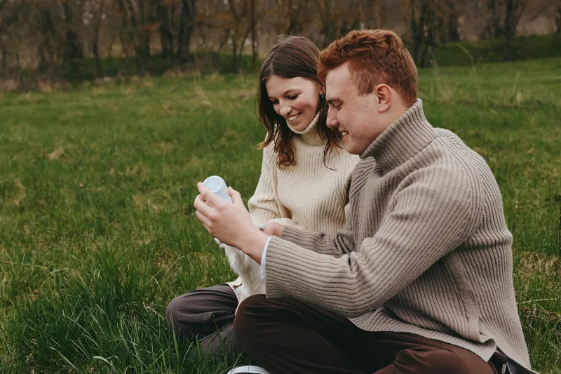 Smiling couple outdoors gently holds a goat kid in a grassy field, celebrating a warm, natural moment of connection, care, and rural lifestyle, with soft daylight highlighting affection and playful