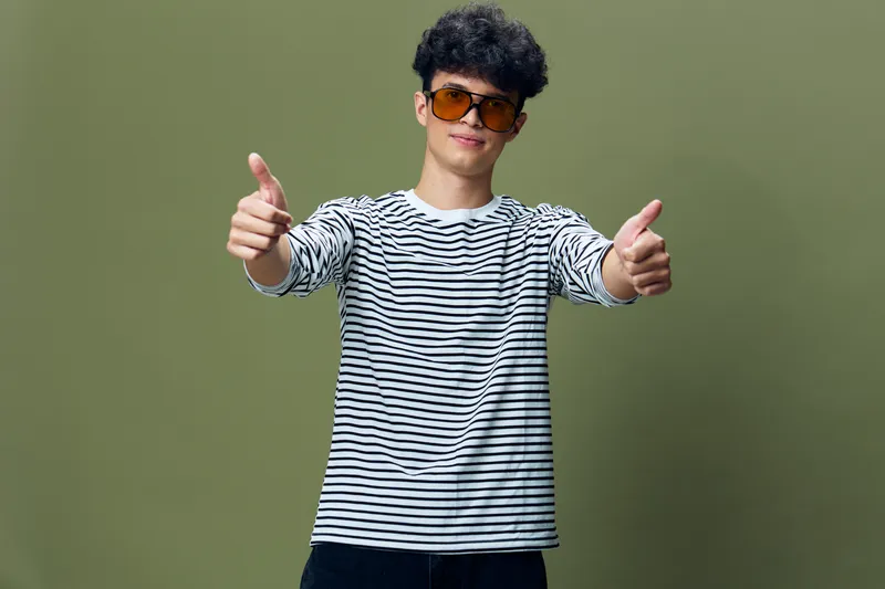 Young man wearing sunglasses and striped shirt shows thumbs up gesture with both hands against olive green background, expressing approval and positivity.