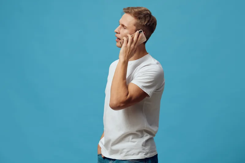 Man talking on mobile phone isolated on plain solid blue background. Studio portrait of a young casual man with short hair wearing white t-shirt using cellphone. People lifestyle concept, communicatio