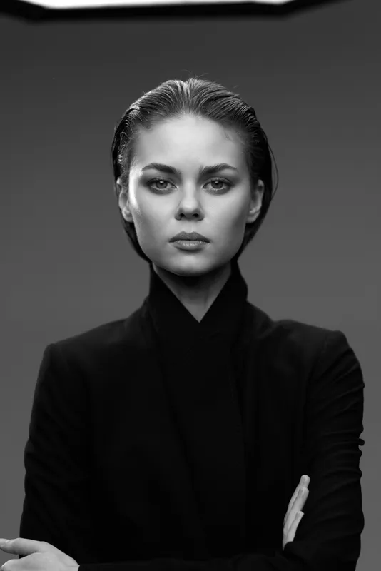 Black and white portrait of serious woman in black clothing with intense gaze and crossed arms.