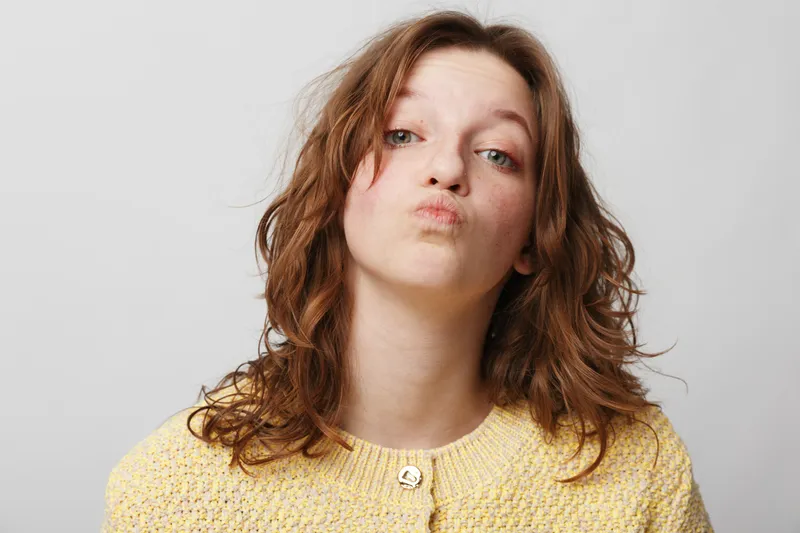 Young woman with curly brown hair wearing yellow sweater makes funny face with pursed lips against plain white background. Concept of humor, expression, casual style, and personality.