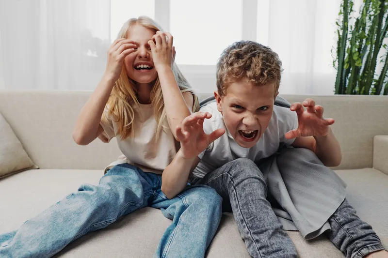 Two playful children sitting on a couch making silly faces and gestures in a bright living room. Concept of childhood fun, family moments, and playful emotions at home.