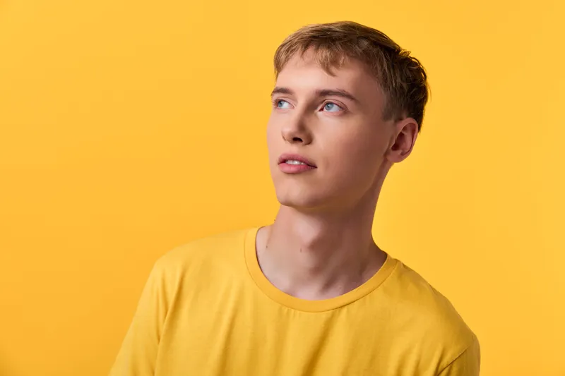 Yellow background with a man in a bright yellow shirt, looking upward and to the side, conveying curiosity, style and casual confidence in a clean studio setting