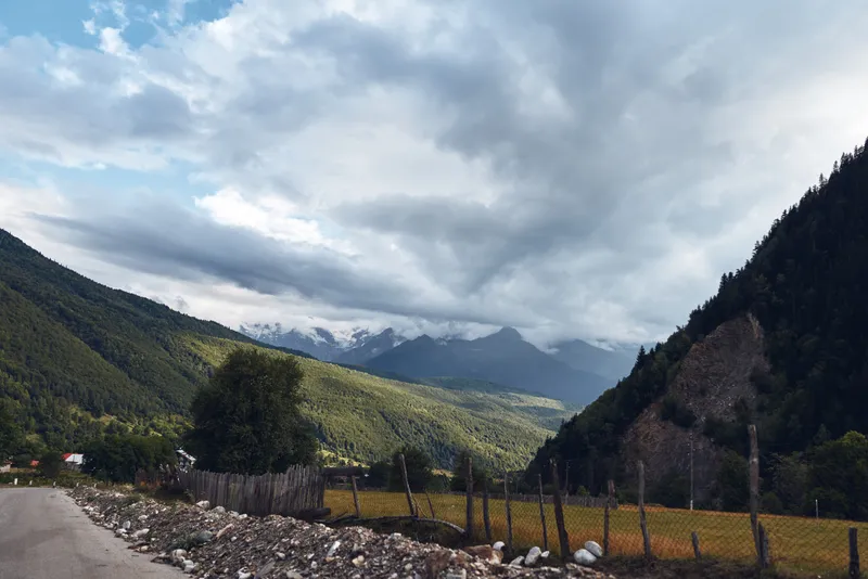 Majestic mountains flank a winding road through a tranquil valley, forming an expansive landscape scene under moody dramatic clouds overhead and open sky.