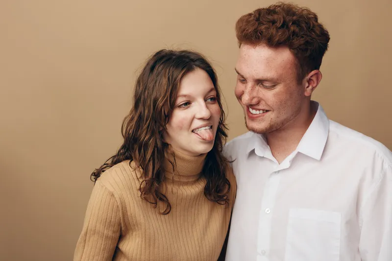Young couple having fun together, woman playfully sticking out tongue while man smiles. Casual clothing and warm beige background create relaxed, joyful mood of friendship and affection.