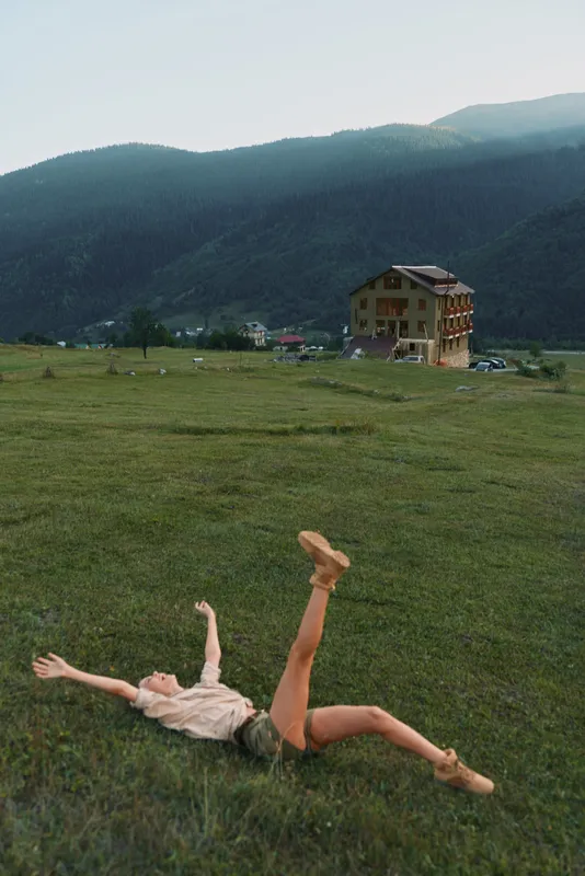 A young woman lies on a grassy field with one leg raised, expressing carefree energy as a bright mountain landscape and a distant building create a dramatic backdrop.