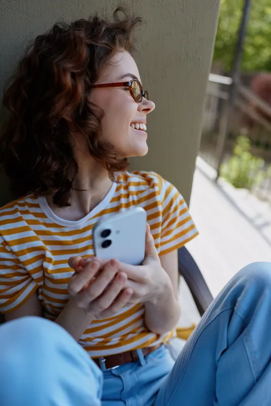 Woman outdoors, smiling, seated, holding a phone, striped shirt, glasses, sunlit moment, casual pose, relaxed lifestyle, candid portrait against a bright outdoor backdrop
