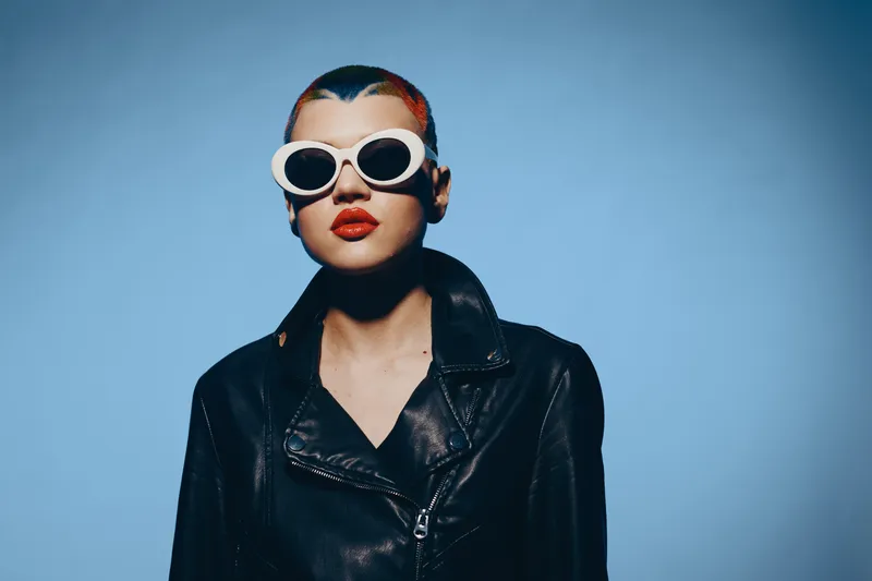 Woman with short colorful hair wearing white sunglasses and a black leather jacket against a blue background, showcasing bold fashion and confident attitude.
