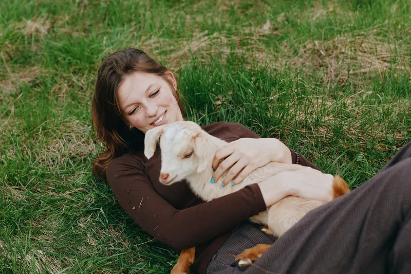Relaxed woman lying on grass with a goat kid cuddling nearby, an intimate outdoor lifestyle moment showing affection, calm breeze, soft light, natural setting, warm bond, candid atmosphere, gentle