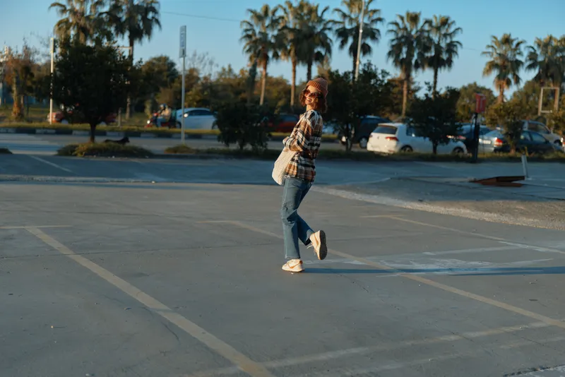 woman walking in parking with sunglasses and bag, casual outfit and denim sneakers under golden hour glow, candid lifestyle, authenticity and emotional storytelling in urban palms scene