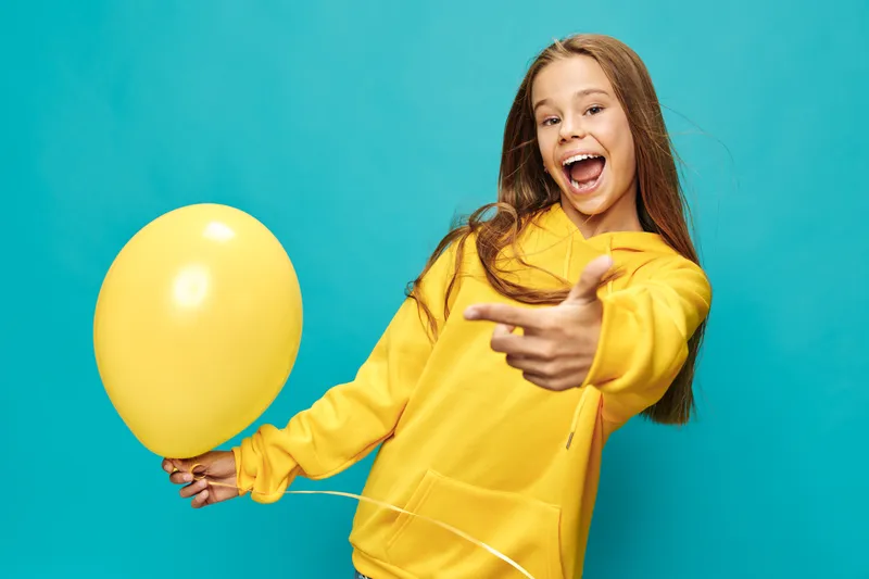 Girl in yellow hoodie holding balloon and smiling with open mouth on blue background. Youthful happy child with long hair and casual clothing celebrating with party balloon.