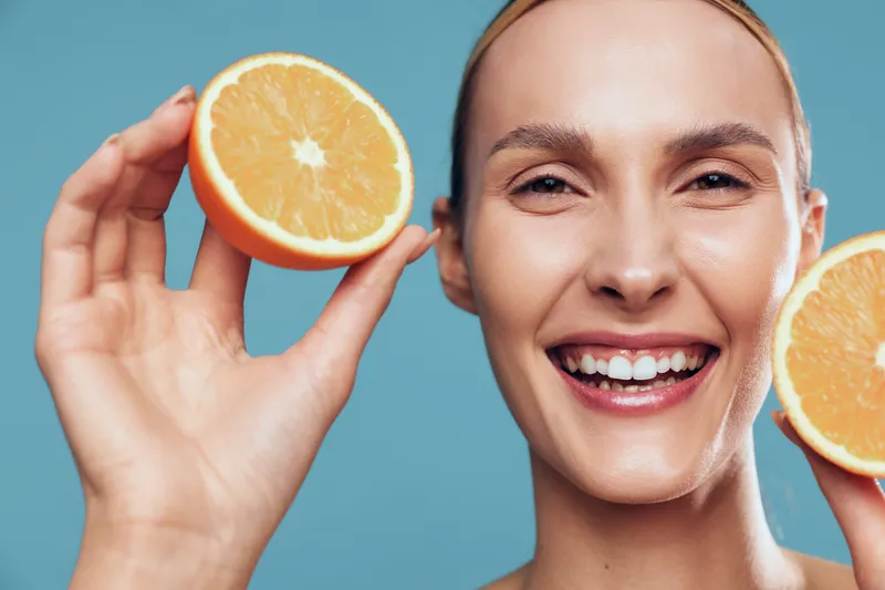 Young woman with clear skin smiles joyfully while holding two halves of a fresh orange fruit against a plain blue background in a bright studio setting.