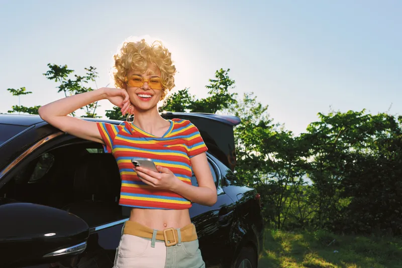Cheerful woman outdoors, smiling beside a car, striped crop top, soft sunlight and relaxed casual vibe, retro fashion colors, lush greenery, sunny summer moment