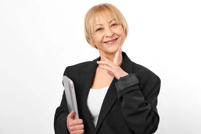 Woman senior in black blazer, smiling in studio, holding smartphone, confident pose, gentle hand gesture, clean white background, soft light, neutral colors