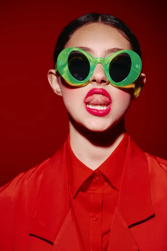 Fashion model in bright red outfit and large green sunglasses poses expressively with tongue out smeared lipstick. Studio shot with intense lighting on a red background. Beauty creative concept.