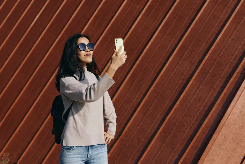 Selfie of a casual woman wearing sunglasses and backpack, holding a smartphone against a warm wooden wall, urban outdoor style, sunny day, candid lifestyle moment captured for social media
