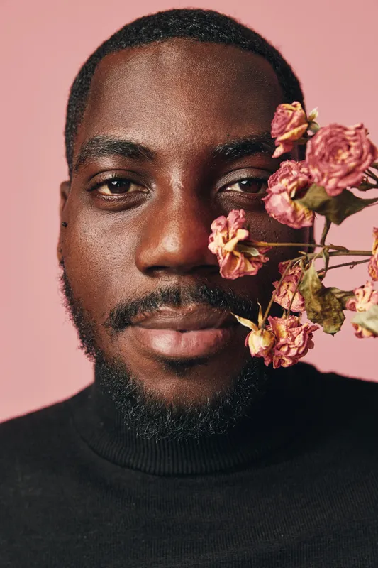 African man portrait with flowers close to face in creative conceptual visual style on pink background. Black male model with beard wearing black turtleneck for artistic expression.