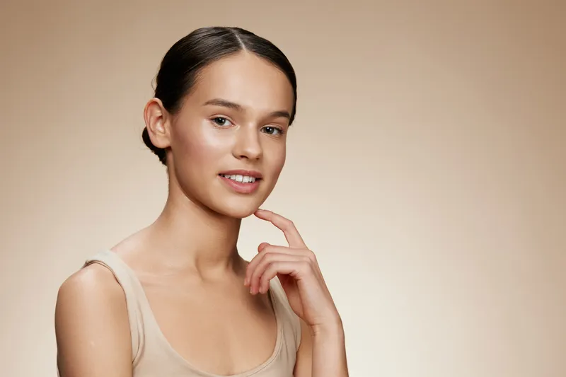 Young female model with a natural, radiant smile, wearing minimal makeup against a soft beige background, exuding confidence and elegance