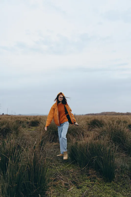 Woman lifestyle walking field outdoors nature autumn countryside, full body portrait on grassy path, warm orange jacket and scarf, relaxed smile, soft cloudy sky background, peaceful mood