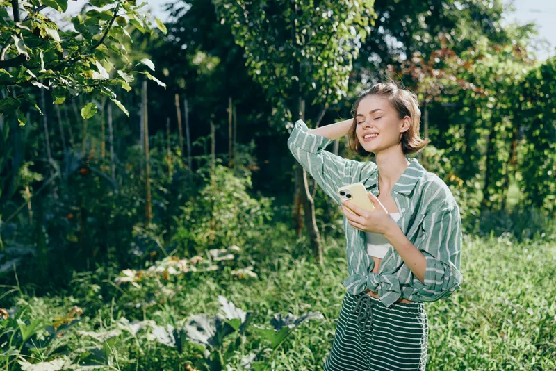 A joyful young woman stands in a sunlit garden, smiling while holding a phone and a plant.