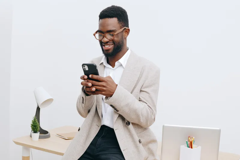 Smiling happy cheerful young African American man wearing glasses, beige sweater, white shirt, using smartphone in modern office space with white wall, minimal decor, and bookshelf, lifestyle concept.