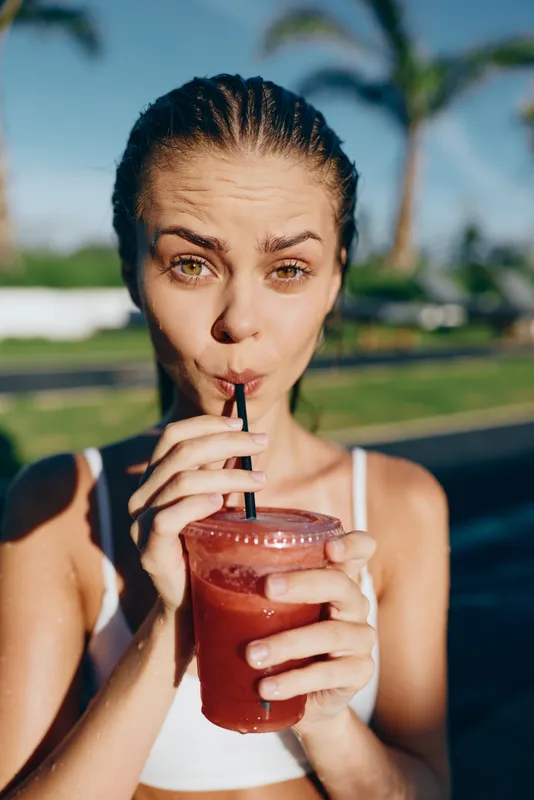 A woman drinks a red smoothie outdoors on a sunny day, poolside setting, wearing a white top, straw in hand. A casual, healthy lifestyle moment captured for wellness and leisure themes.