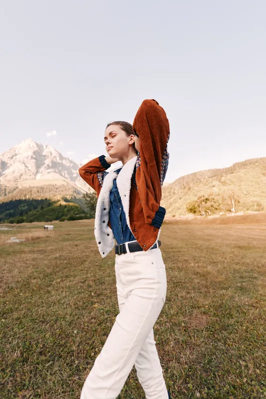 Woman fashion mountain outdoors autumn portrait of a young model standing in a scenic meadow, wearing cozy sweater and white trousers, posing confidently with sunlight and serene landscape.