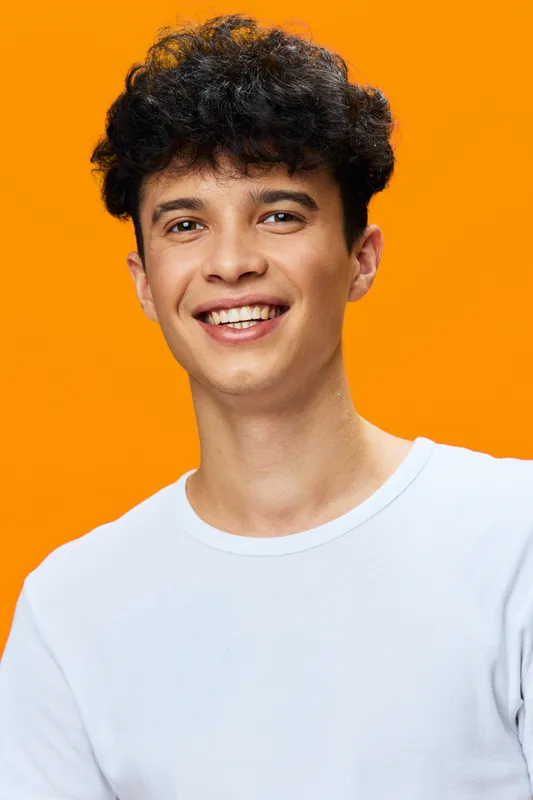 smiling man with curly hair wearing a white shirt stands against bright orange background, showing youthful energy and positive attitude in casual portrait style