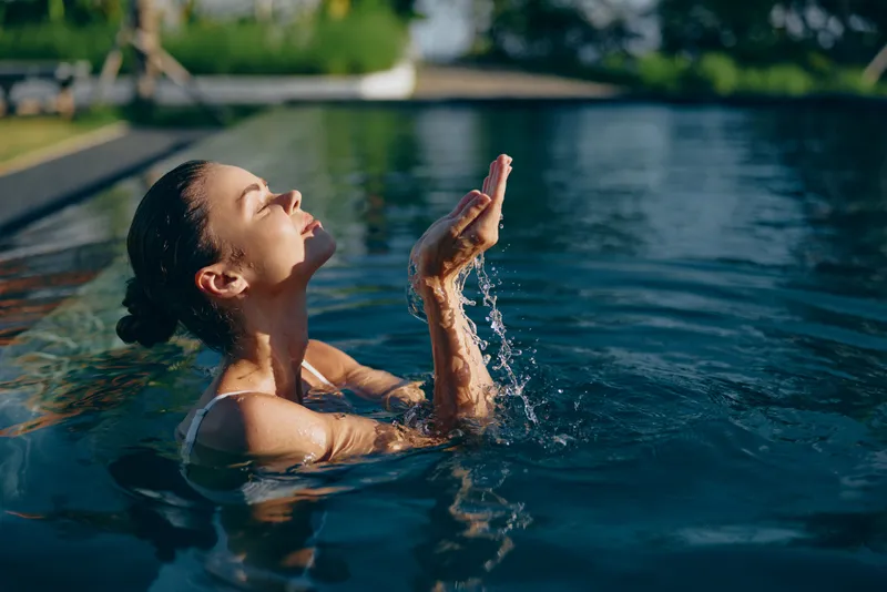 Relaxed woman bathes in a sunlit pool, cupping water with both hands, enjoying the refreshing moment, outdoor poolside scene, summer leisure, tranquil mood, blue water glow, candid lifestyle