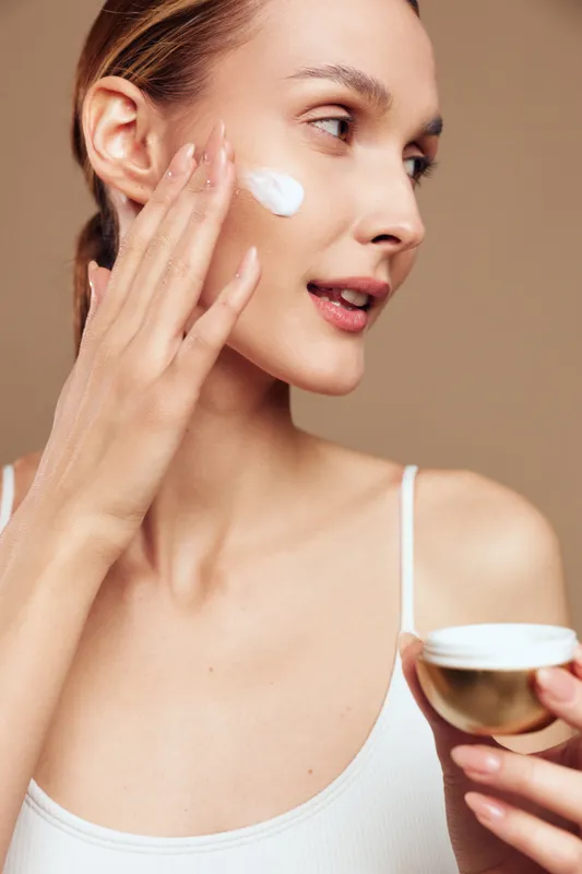 Young woman applies moisturizing cream on her face with fingers. Skincare routine concept with natural skin, beauty, and self-care on beige background in soft light.