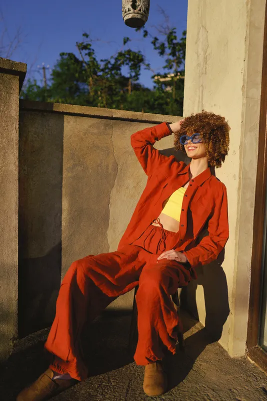 Young woman wearing a red suit lounges on a sunlit balcony, posing casually with sunglasses and a relaxed smile, emanating confident, carefree lifestyle vibes outdoors