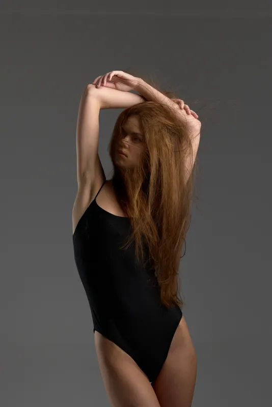 Young woman with long red hair posing in black swimsuit against gray background, expressing elegance and confidence.
