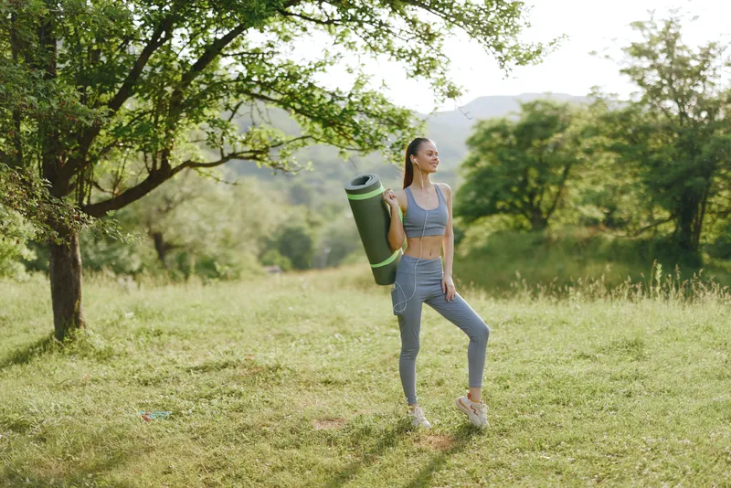 Young woman in athletic wear holding yoga mat, smiling in a lush green outdoor setting, embodying fitness and a healthy lifestyle