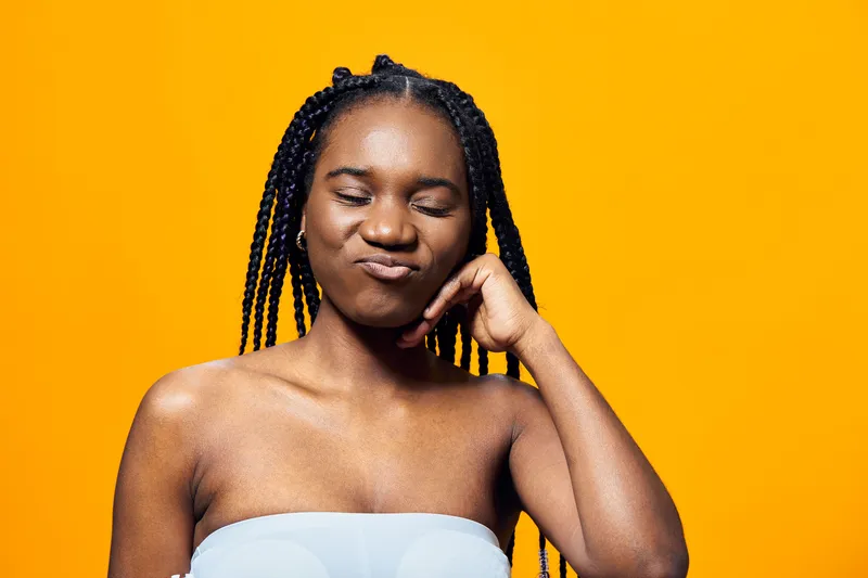 Young African woman with braided hair expressing playful frustration against a vibrant yellow background, showcasing summer vibes and confident emotion