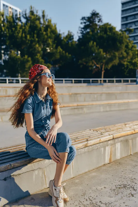 Young woman with long red hair sitting outdoors wearing sunglasses and denim outfit, enjoying sunny day with urban background and relaxed mood.