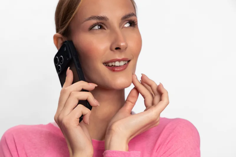 Young woman in pink sweater holds smartphone near ear, smiling and looking away. Bright studio portrait with natural makeup, concept of communication, technology, and positive emotions.