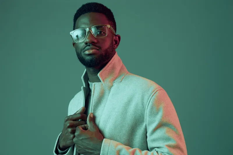 Stylish African man in a modern coat, showcasing confidence against a green background with soft lighting, perfect for fashion and lifestyle concepts