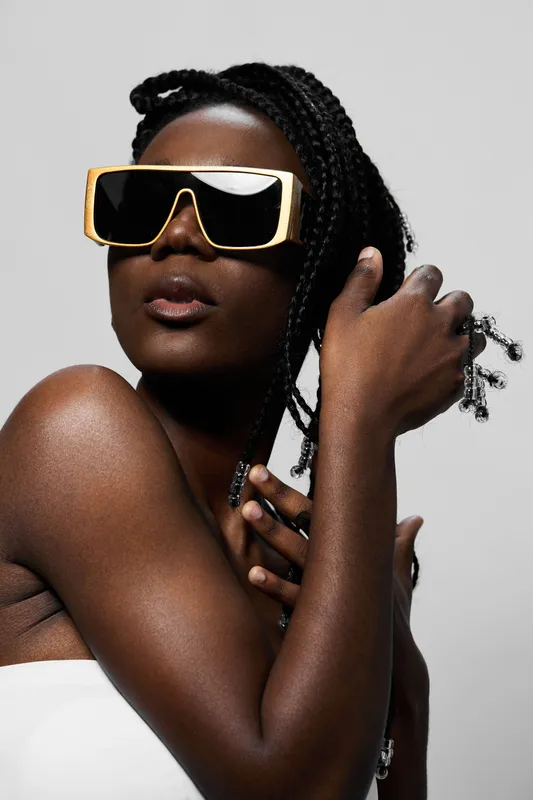 Stylish woman with braided hair and oversized sunglasses poses confidently against a light background, embodying modern fashion trends