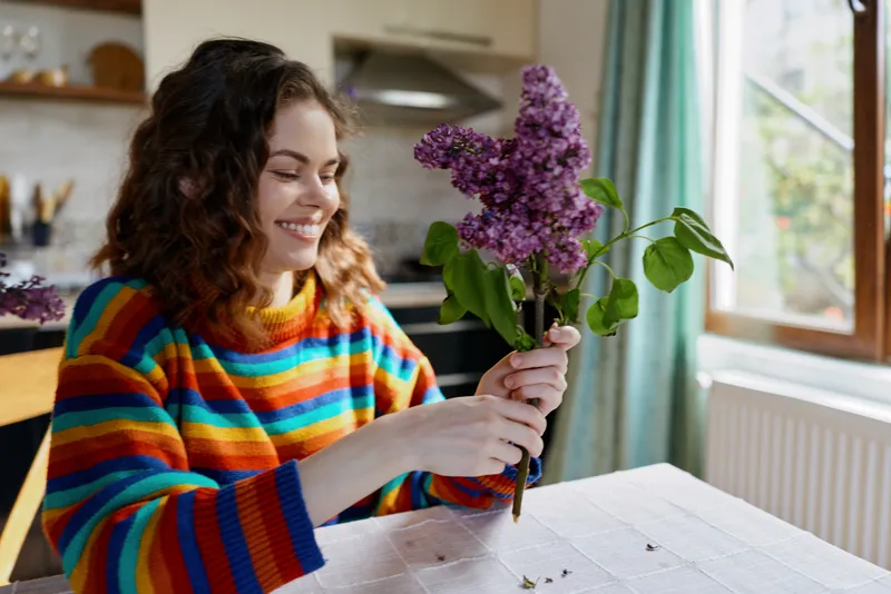 A smiling woman in a colorful striped sweater holds a bouquet of purple flowers at a sunny kitchen table, capturing a cozy home moment and hobby indoors with bright kitchen light.