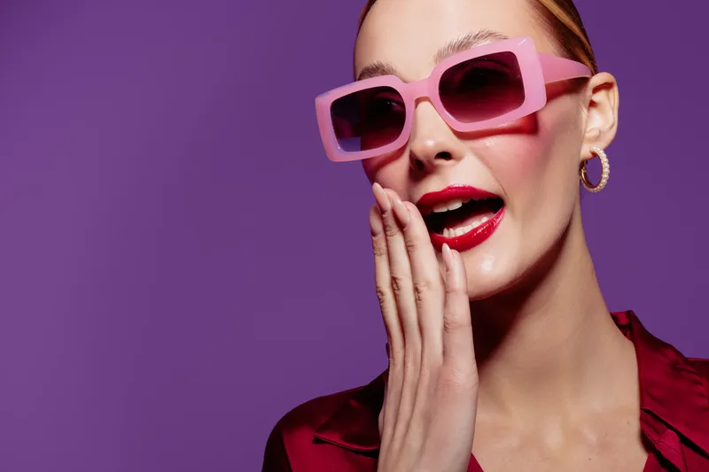 Stylish woman in pink sunglasses and red lipstick poses with hand near mouth on purple background. Concept of fashion, beauty, glamour, and confident female model portrait.